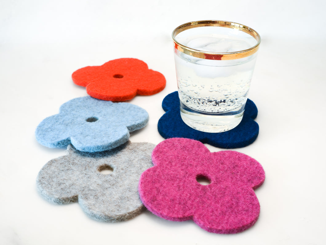 White background with five, colorful flower coasters laid out in a circle in red, light blue, pink, navy and grey.  A gold rimmed glass with a fizzy drink is sitting on the blue coaster. The coasters are flower shaped and made of 1/4