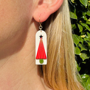 Model wearing a white arched earring with  red tree with metallic gold lines and green trunk and star cut out at top of tree by Bird of Virtue