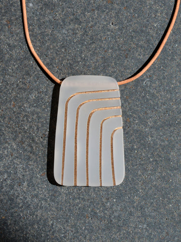 Rectangular gray pendant with curved gold lines on leather cord necklace, photographed on grey stone surface