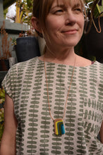Woman wearing geometric print top with colorful layered pendant necklace, smiling outdoors with plants in background