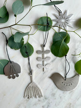 A styled flat-lay of four handcrafted metal  hanging ornaments on a white marble surface, surrounded by green eucalyptus leaves. The ornaments feature organic shapes including a bird, a leafy sprig, a flower, and a star, in brushed stainless steel.