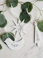 Three white metal Christmas ornaments shaped like a star, a dove, and a Christmas tree, hanging from white strings on green eucalyptus branches over a light marble surface.