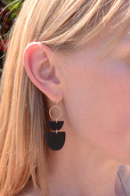 A close-up side profile of a woman with blonde hair wearing a pair of black geometric dangle earrings with gold-tone circular accents, set against a blurred outdoor background of greenery and red flowers.
