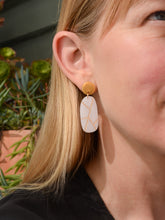 White oval earring with gold geometric lines worn by a blonde woman in a black top with plants in background. Made by Bird of Virtue - San Francisco, CA