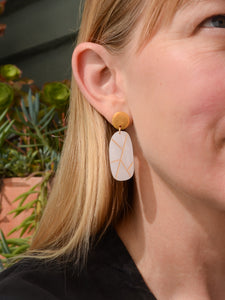 White oval earring with gold geometric lines worn by a blonde woman in a black top with plants in background. Made by Bird of Virtue - San Francisco, CA