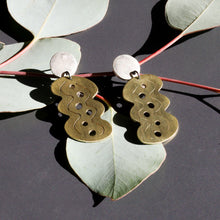 Brass earrings with wavy patterns on a dark surface with green leaves.