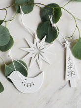 Three white steel Christmas ornaments shaped like a star, a dove, and a Christmas tree, hanging from white strings on green eucalyptus branches over a light marble surface.