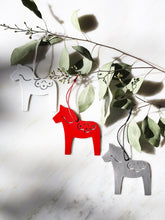 Three metal Swedish Dala horse-shaped ornaments in white, red, and brushed stainless, hanging from a leafy branch against a white marble background.