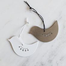 Two bird-shaped hanging ornaments on a white marble surface, one white with a white hanging string and one brushed stainless steel with a black hanging string.