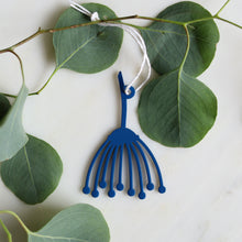 A blue decorative hanging ornament shaped like a stylized Queen Anne's Lace flower, suspended from a white string and styled among green eucalyptus leaves on a white marble surface.