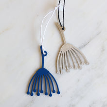 Two small decorative Queen Annes Lace floral ornaments displayed on a white marble surface, one painted blue with a white hanging cord and one brushed stainless steel with a black hanging cord.