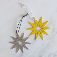 Two star-shaped metal ornaments on a white marble surface, one in bright yellow with a white hanging cord and one in brushed stainless steel with a dark gray hanging cord.