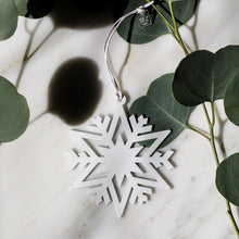 A white snowflake-shaped hanging ornament with a white string, styled on a white marble surface with green eucalyptus leaves.