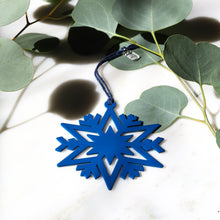 A blue snowflake-shaped hanging ornament with a dark blue cord, styled on a white marble surface with green eucalyptus leaves.