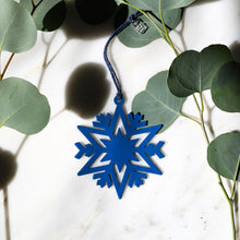 A blue laser-cut snowflake ornament hanging from a dark blue string, styled on a white marble surface with green eucalyptus leaves.