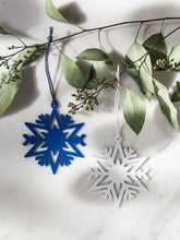 Two metal snowflake ornaments, one in blue and one white, hanging from green eucalyptus branches on a white marble surface.