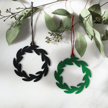 Two circular wreath-shaped hanging ornaments, one black and one green, displayed on a white marble surface with eucalyptus branches. The black ornament hangs from a dark cord, and the green ornament hangs from a burgundy cord.