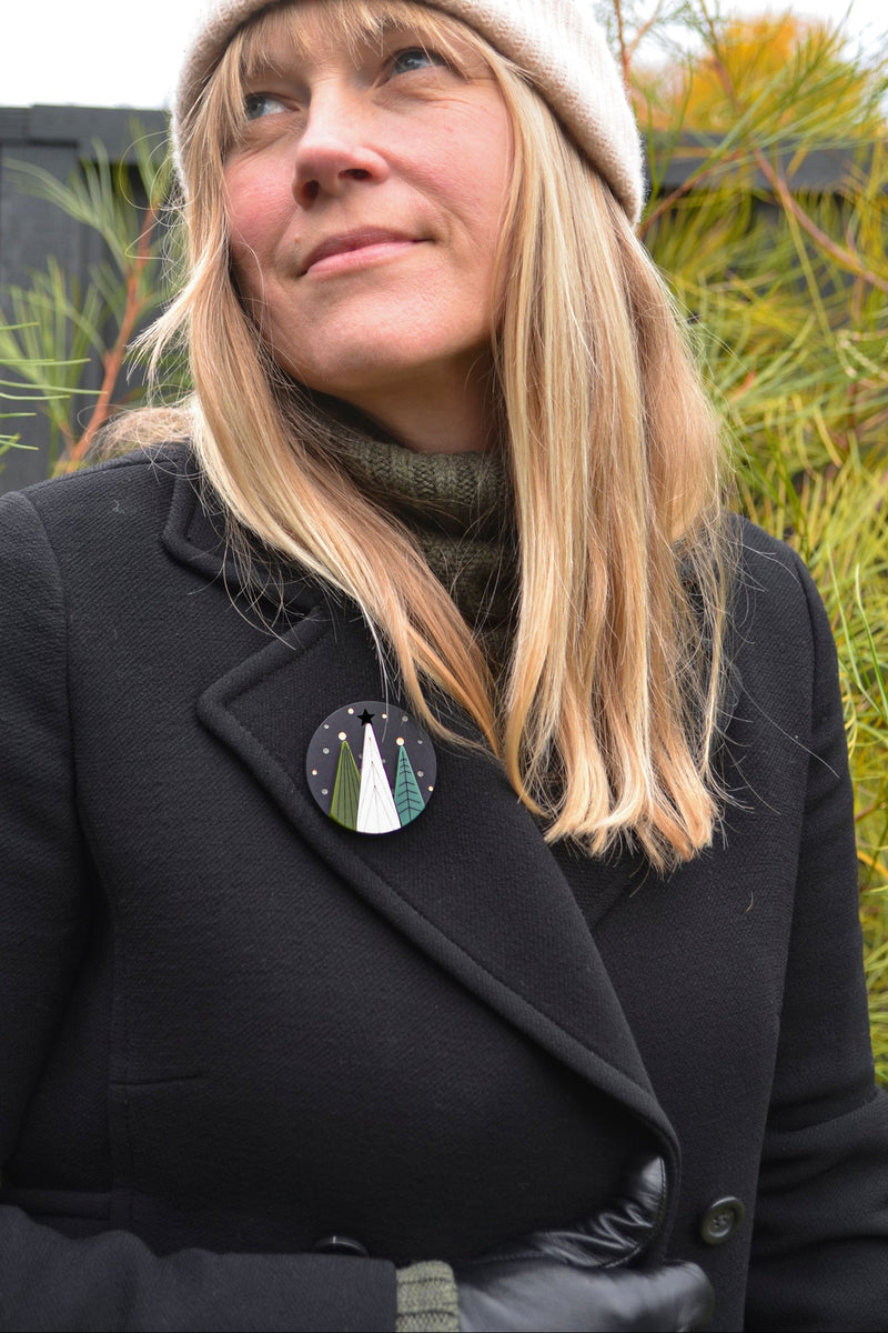 Person wearing a black coat with a tree-themed round brooch, standing outdoors.