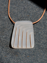 Rectangular gray pendant with curved gold lines on leather cord necklace, photographed on grey stone