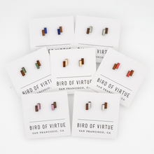 Seven pairs of geometric wooden stud earrings from Bird of Virtue featuring rectangular blocks in various colors including blue, green, red, pink, and white.