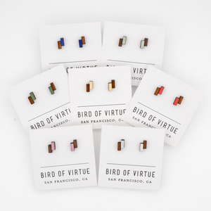 Seven pairs of geometric wooden stud earrings from Bird of Virtue featuring rectangular blocks in various colors including blue, green, red, pink, and white.