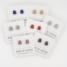 Bird of Virtue's rainbow arch stud earrings in seven colors displayed on branded cards from San Francisco. Wooden half-circle design with rainbow arch patterns.