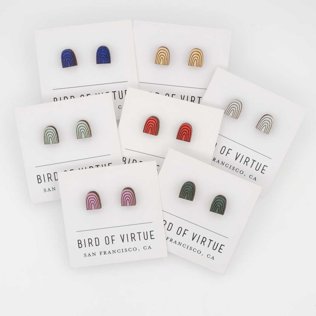 Bird of Virtue's rainbow arch stud earrings in seven colors displayed on branded cards from San Francisco. Wooden half-circle design with rainbow arch patterns.