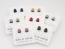 Bird of Virtue's rainbow arch stud earrings in seven colors displayed on branded cards from San Francisco. Wooden half-circle design with rainbow arch patterns.