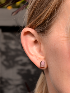 Close-up of an ear wearing a natural wood stud earring painted orchid purple with engraved rainbow arch design, showcasing its minimalist style on fair skin.