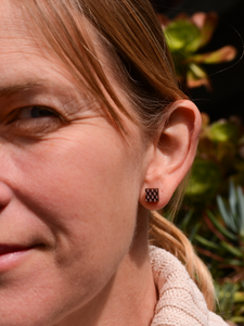 Close-up of a woman wearing a dark brown wooden checkerboard pattern stud earring from Bird of Virtue against a green plant background.