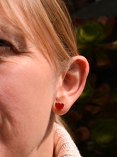 A close-up of a woman's face, showcasing a small red flower-shaped stud earring with a delicate black floral design, with greenery in the background.