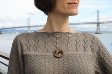 Person wearing a patterned sweater with a necklace featuring a wooden pendant, standing in front of a bridge.