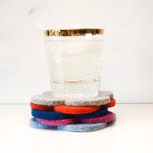 White background with five colorful wool coasters stacked with a gold-rimmed glass with a fizzy drink sitting on top of it. The coaster colors are light blue, pink, navy, red and grey. The coasters are made of 1/4" thick wool felt and have a slight textured appearance.