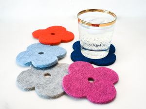 White background with five, colorful flower coasters laid out in a circle in red, light blue, pink, navy and grey.  A gold rimmed glass with a fizzy drink is sitting on the blue coaster. The coasters are flower shaped and made of 1/4" thick wool. 