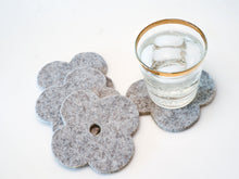 White background with four grey flower coasters laid out and a gold-rimmed glass filled with ice and a fizzy drink sitting on one of the coasters for scale. The coasters are made of 1/4" thick wool design felt and have a slight textured appearance.