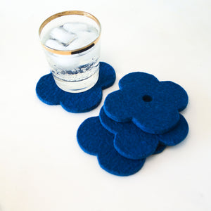 White background with four navy blue flower coasters laid out and a gold-rimmed glass filled with ice and a fizzy drink sitting on one of the coasters for scale. The coasters are made of 1/4" thick wool design felt and have a slight textured appearance.