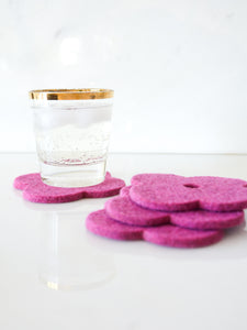 White background with four pink flower coasters laid out and a gold-rimmed glass filled with ice and a fizzy drink sitting on one of the coasters for scale. The coasters are made of 1/4" thick wool design felt and have a slight textured appearance.
