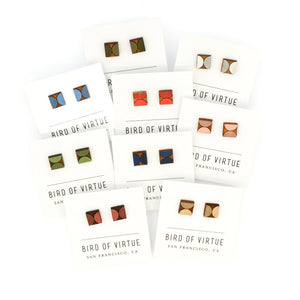Assortment of colorful, wooden square stud earrings with semicircles engraved on them. Displayed on white Bird of Virtue cards against a white background. Colors are silver, moss, gold, leaf, poppy, midnight, dogwood, rouge, sky blue. Made by Bird of Virtue